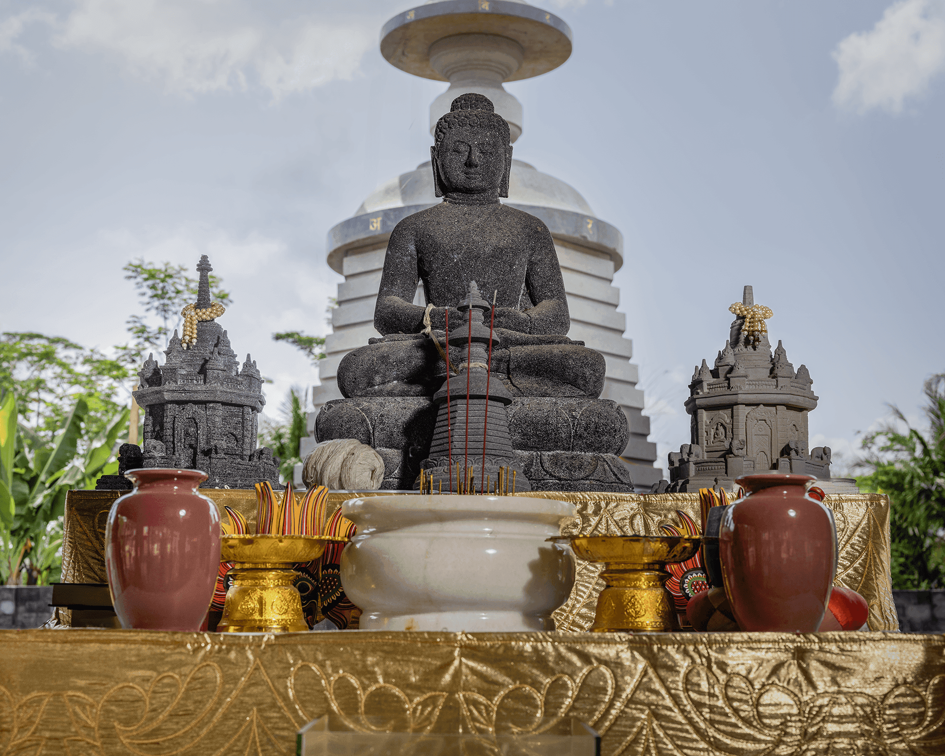 Patung Buddha duduk di Pusdiklat Catra Jinadhammo, yang di luar ruangan dan dikelilingi oleh vas serta artefak Buddhis lain.